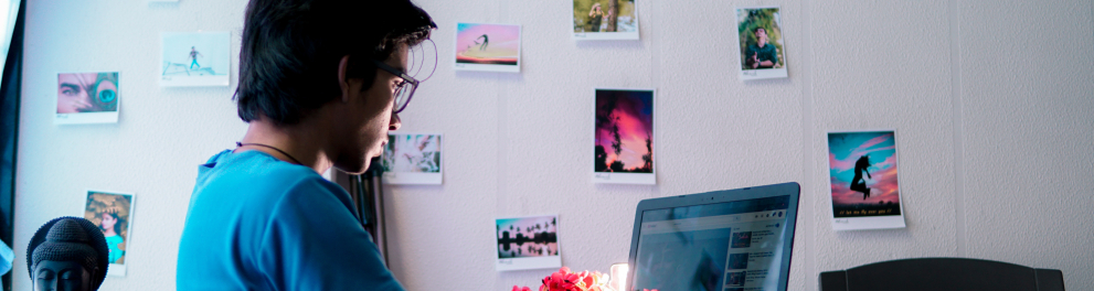 Um jovem, que usa óculos, está sentado diante do computador. A parede, ao fundo, é branca, mas com diversas fotografias coladas nela.