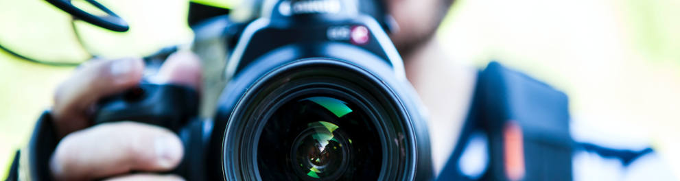 Imagem focada na lente de uma câmera de filmar que está sendo manuseada por um homem branco ao fundo