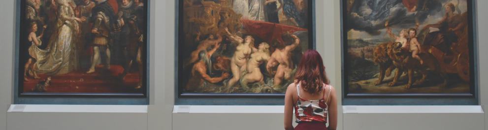 Imagem de uma mulher sentada em um banco, de costas para quem bateu a foto e de frente para três quadros grandes. Claramente ela está em um museu