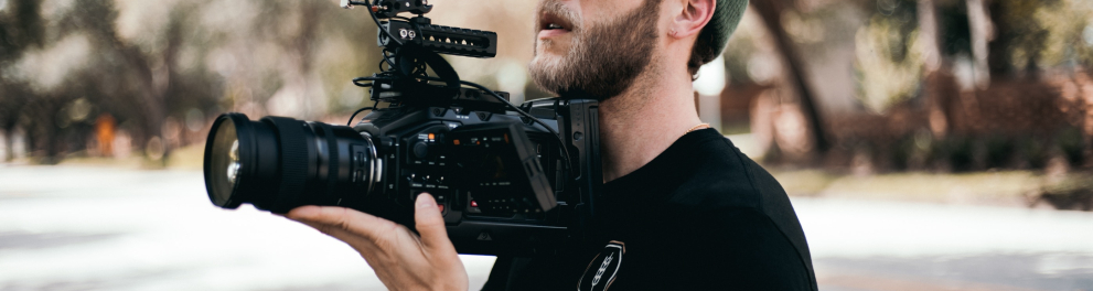 Um homem branco e barbudo está segurando uma câmera de filmar em um ambiente ao ar livre, na rua